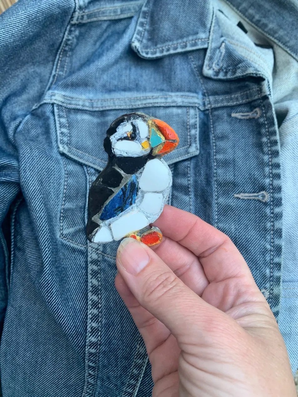 Detailed mosaic puffin brooch held against a denim jacket, in black, white, orange and blue