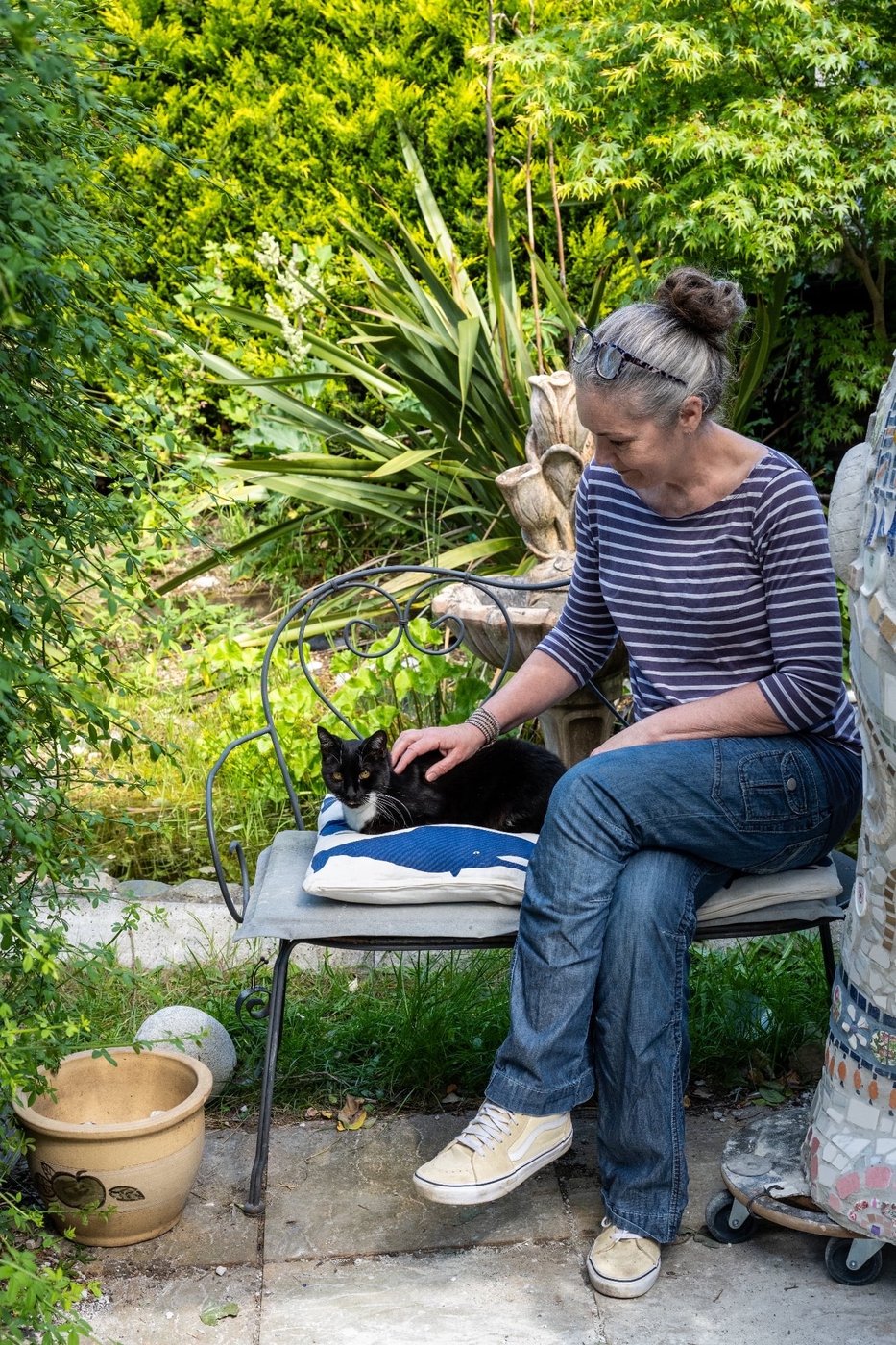 Sarah Delgado Barter in her Suffolk garden, sitting with her cat beside one of her mosaic sculptures