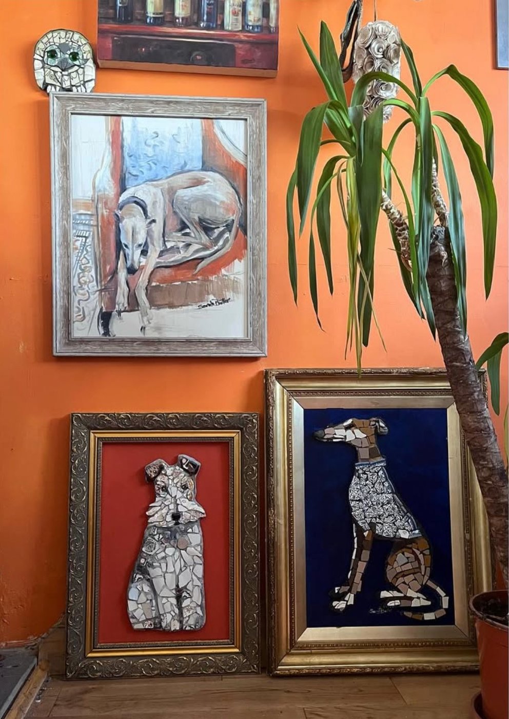 Mosaic whippet portraits in gilt frames beside a painting of a sleeping greyhound